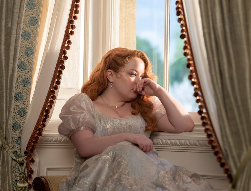 Penelope Featherington sitting down near a window.