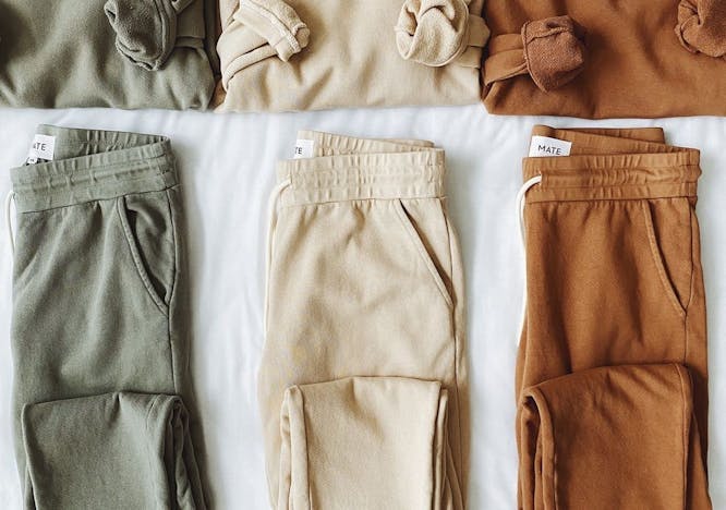Three different sweatshirt and sweatpant sets folded on a bed. The set on the left is green, the set in the middle is beige, and the set on the right is dark orange in color. 