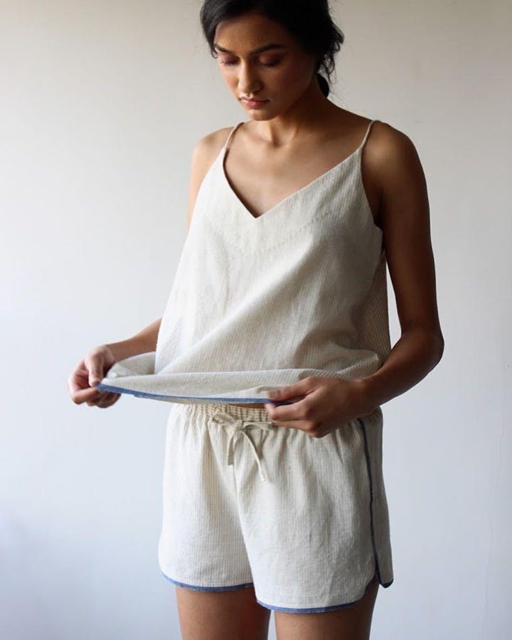 Woman with brown hair wearing a beige tank top and matching short sleep set holding the bottom of her shirt and looking at it. 