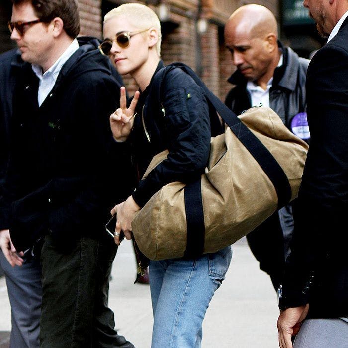 Kristen Stewart with a bleach blonde buzzcut holding a bag with security guards around her.