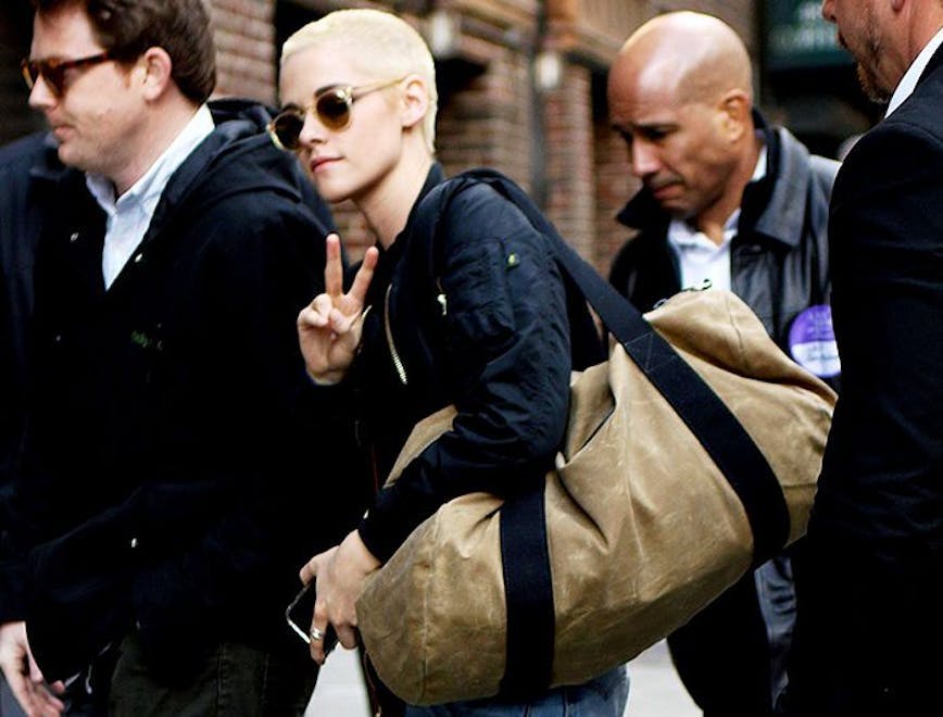 Kristen Stewart with a bleach blonde buzzcut holding a bag with security guards around her.
