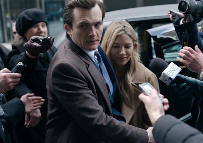 A shot from the new hulu show anatomy of a scandal. Police officers and reporters in the background outside and a man leading a woman in the foreground both in brown coats