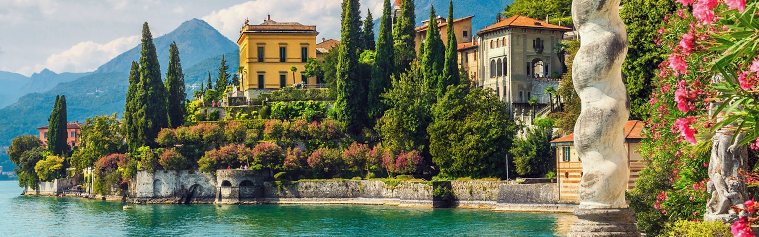 Lake Como Italy with italian houses, the blue lake, and mountains in the background. pink flowers in a bush on the side and a dock in the water with a staircase leading down beside it