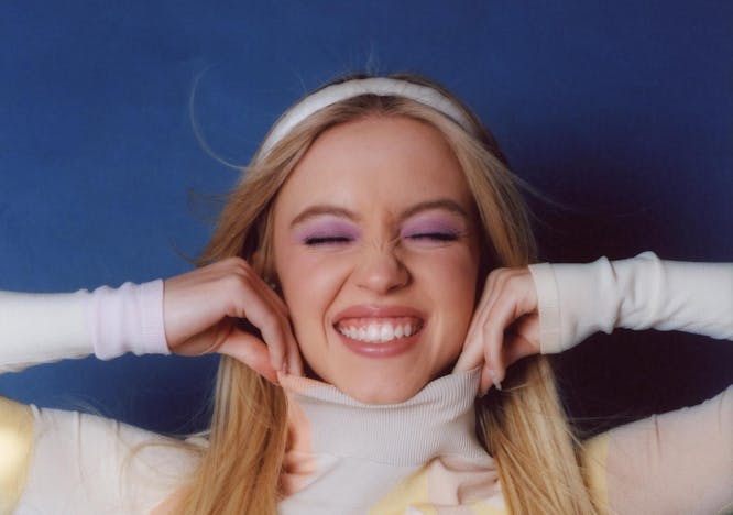 Sydney Sweeney in a multi-colored turtleneck smiling and scrunching her eyes to reveal purple eyeshadow