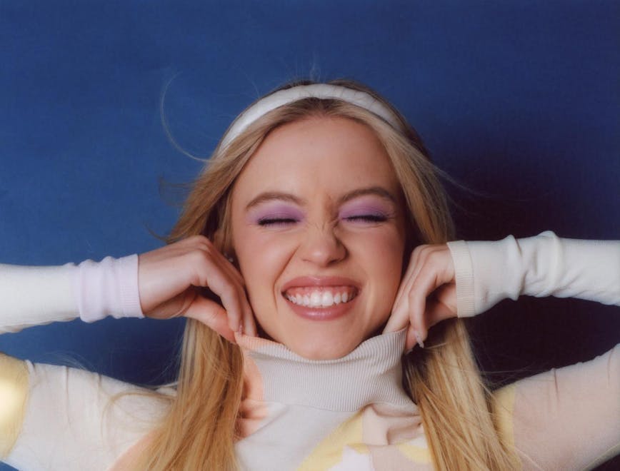Sydney Sweeney in a multi-colored turtleneck smiling and scrunching her eyes to reveal purple eyeshadow