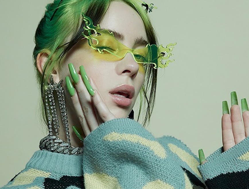 Billie Eilish with green and black hair and long green nails wearing a pair of green flame-shaped sunglasses