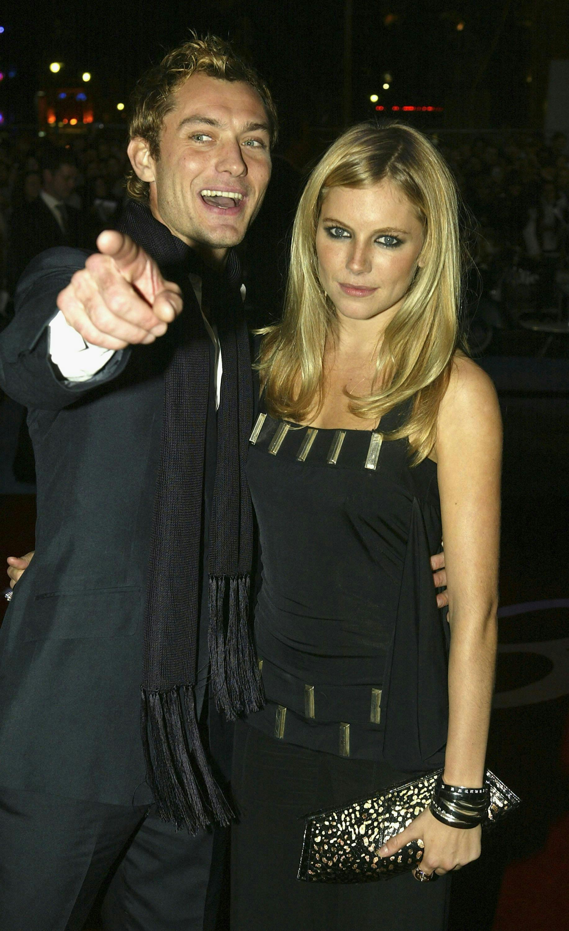 Jude Law and Sienna Miller pose for the camera both wearing all black. Law wears a black scarf and Miller carries a beaded bag.
