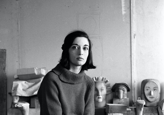 Marisol Sol Escobar sitting in her studio on a small wooden stool. She is surrounded by various sculptures as well as paint cans and wooden barrels. She is wearing a turtleneck sweather, black pants and black boots. She has chin-length black hair, and the picture is in black and white. 