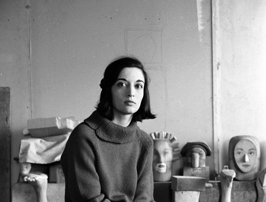 Marisol Sol Escobar sitting in her studio on a small wooden stool. She is surrounded by various sculptures as well as paint cans and wooden barrels. She is wearing a turtleneck sweather, black pants and black boots. She has chin-length black hair, and the picture is in black and white. 