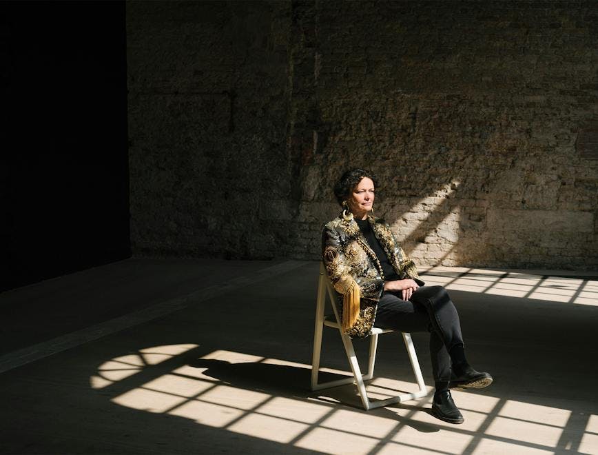 Cecilia Alemani wears orential gold and black Schiaparelli Jacket, black fitted trousers and black boots while sitting on a white foldout chair in the middle of an empty brick walled room. The reflection of large, rounded windows shines on her. 