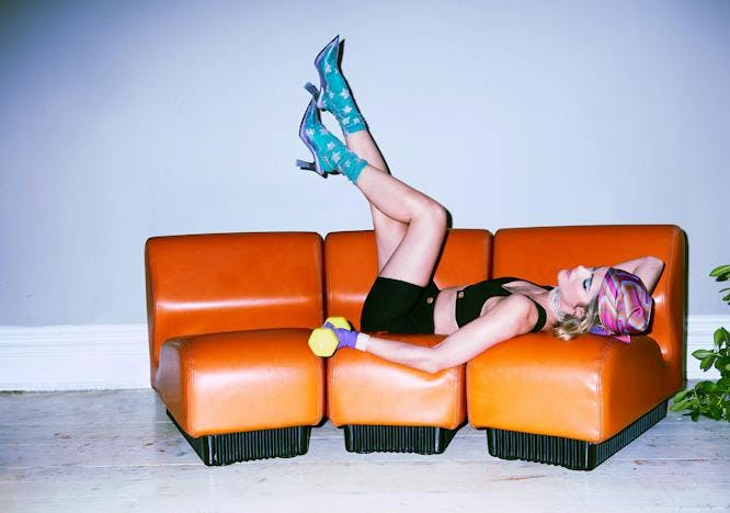 Woman lays on orange couch wearing black sports bra and biker shorts with blue ankle socks and blue Melissa x Larroudé Slingback Heels 