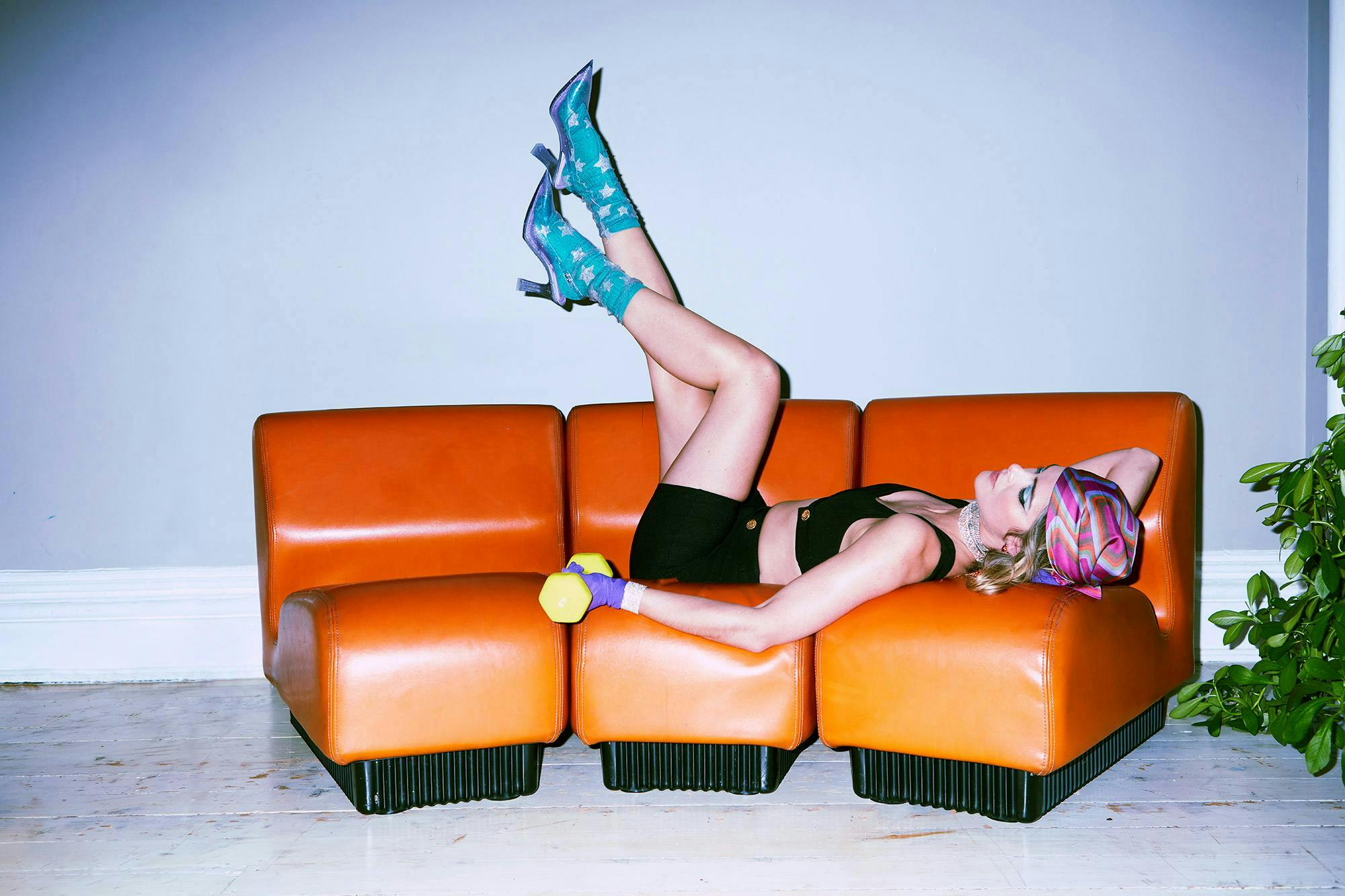 Woman lays on orange couch wearing black sports bra and biker shorts with blue ankle socks and blue Melissa x Larroudé Slingback Heels 