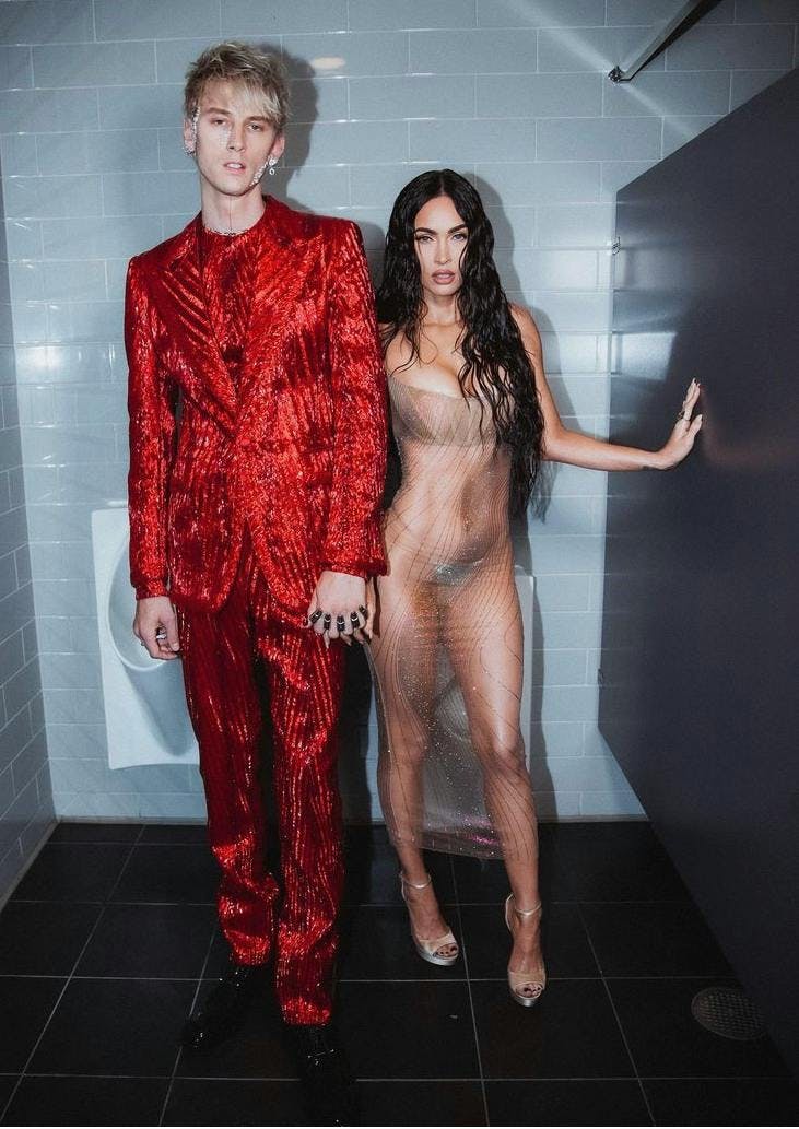 Megan Fox wears a sheer form fitting dress while standing next to fiancé Machine Gun Kelly, who wears a red sparkly suit.