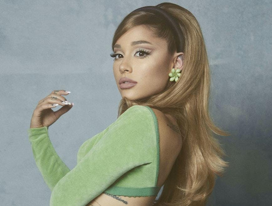 Ariana Grande wears green crop top with matching green flower earrings as she poses in front of a grey back drop.