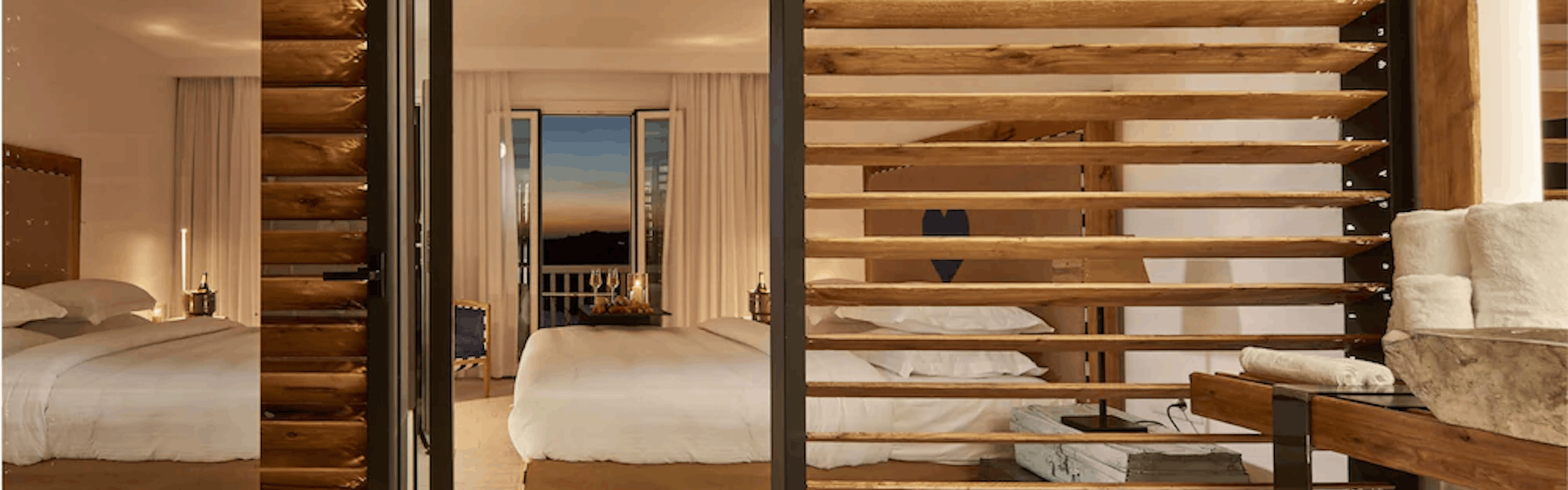 Hotel bed with white sheets, wood details, and a balcony door open with a sunset in background. 