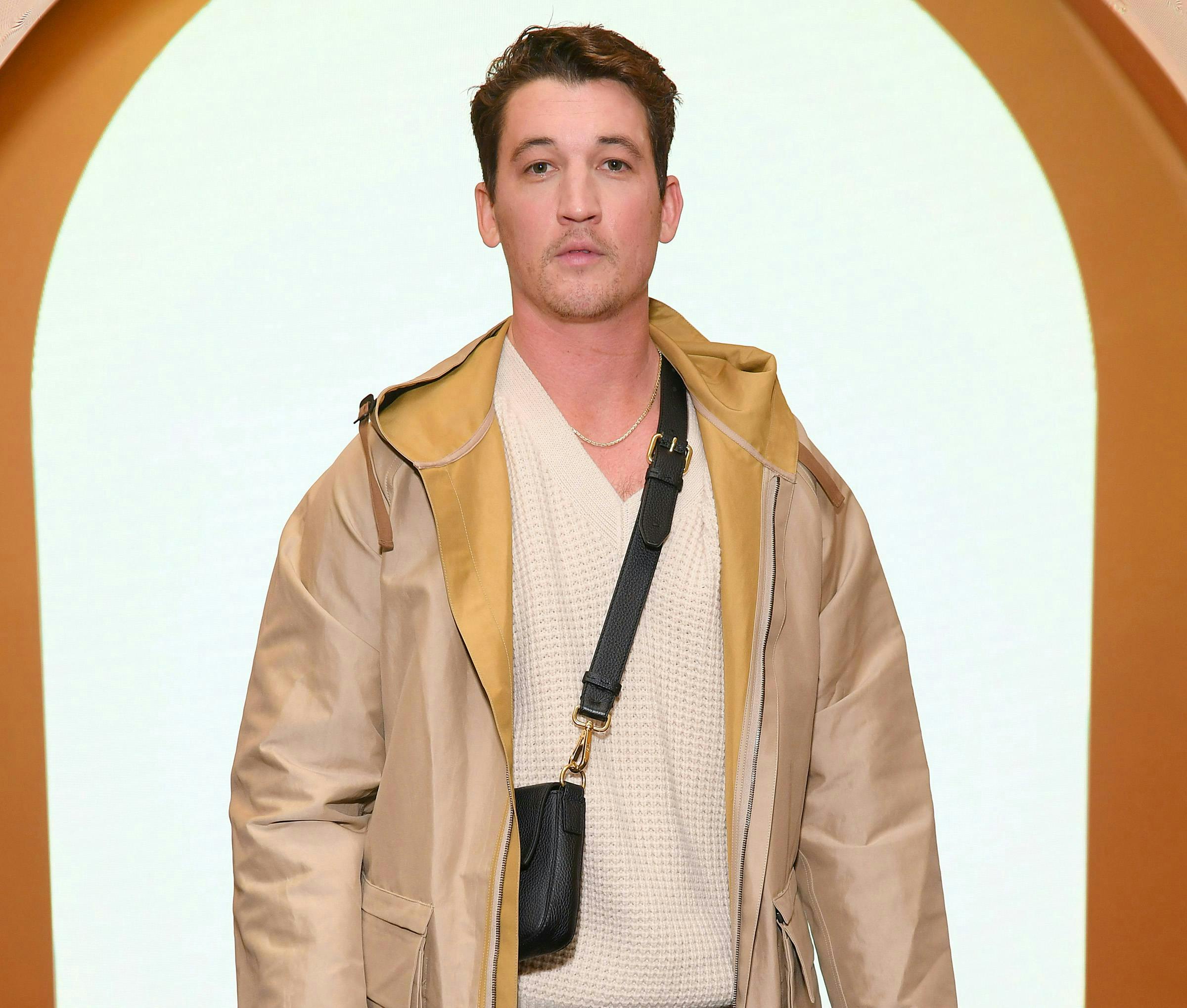 Miles Teller wearing a tan jacket, white sweater and black crossbody bag. 
