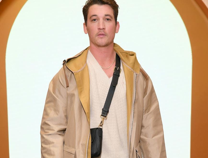 Miles Teller wearing a tan jacket, white sweater and black crossbody bag. 