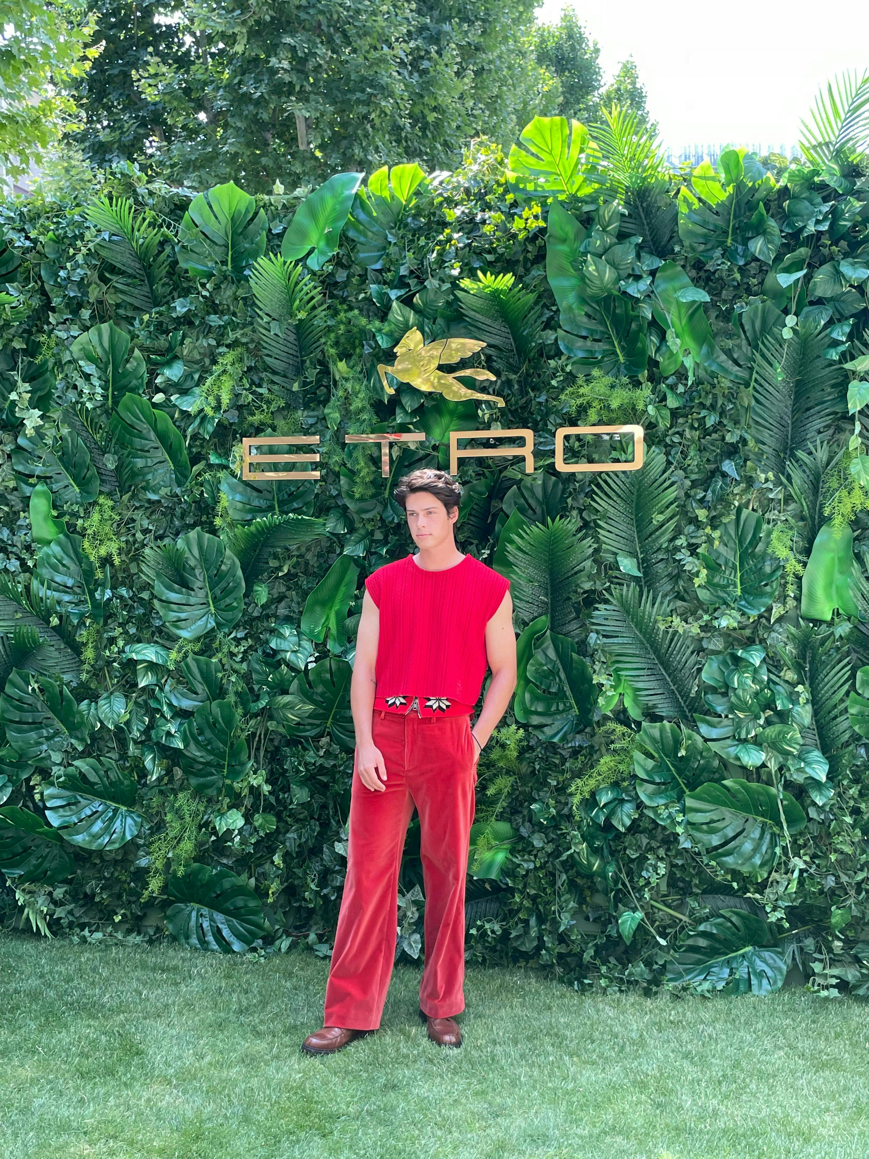 Blake Gray posing in front of a wall of greenery with a gold Etro logo. He is wearing a red sweater vest, red flared pants and brown loafers