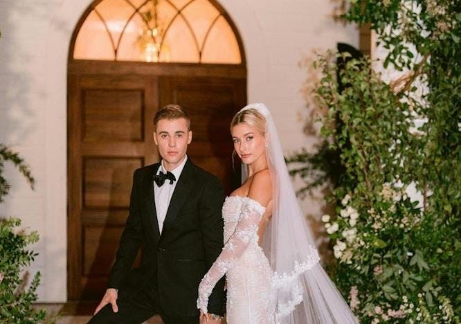 Justin and Hailey Bieber on their wedding day, with Justin wearing a tuxedo and Hailey wearing a white embroidered wedding gown on brick steps