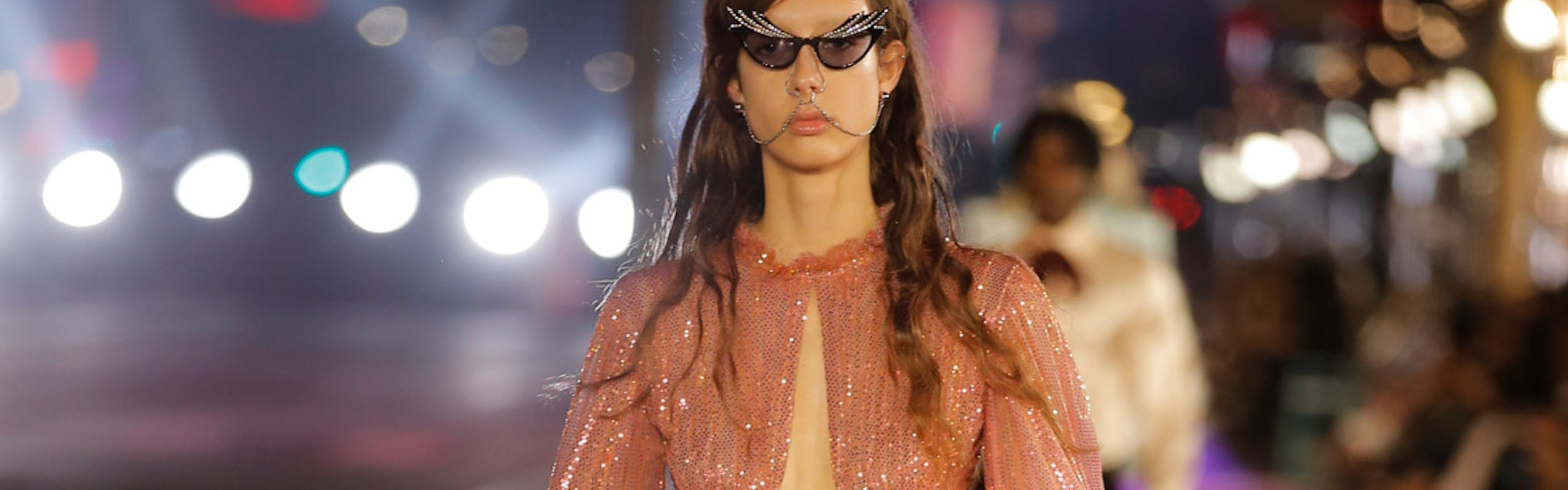 A model in Gucci runway show wearing a salmon shimmery dress with feathery details and black cat eye sunglasses