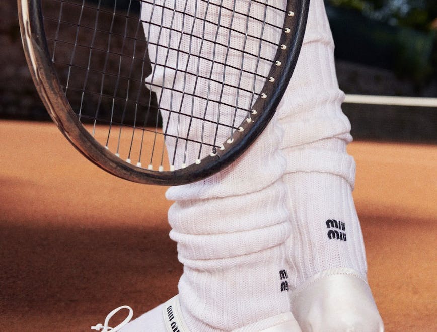 Close up of model wearing white silk Miu Miu ballet flats with leg warmers on a tennis court.