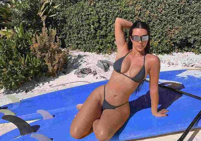 Kim Kardashian poses in a gray bikini on a blue surfboard.