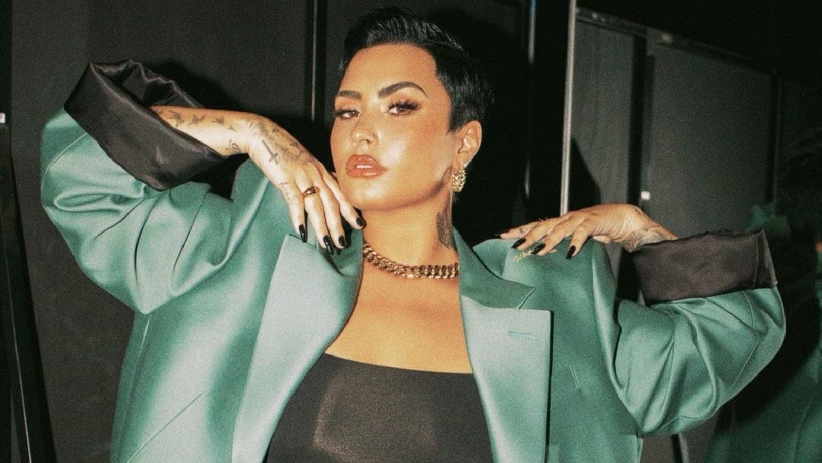 Demi Lovato poses with her arms up, in a green satin suit jacket.
