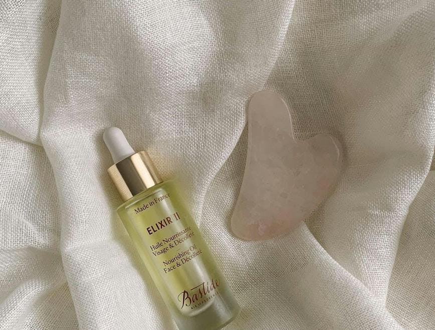 A serum and gua sha lie on a white comforter or sheet.