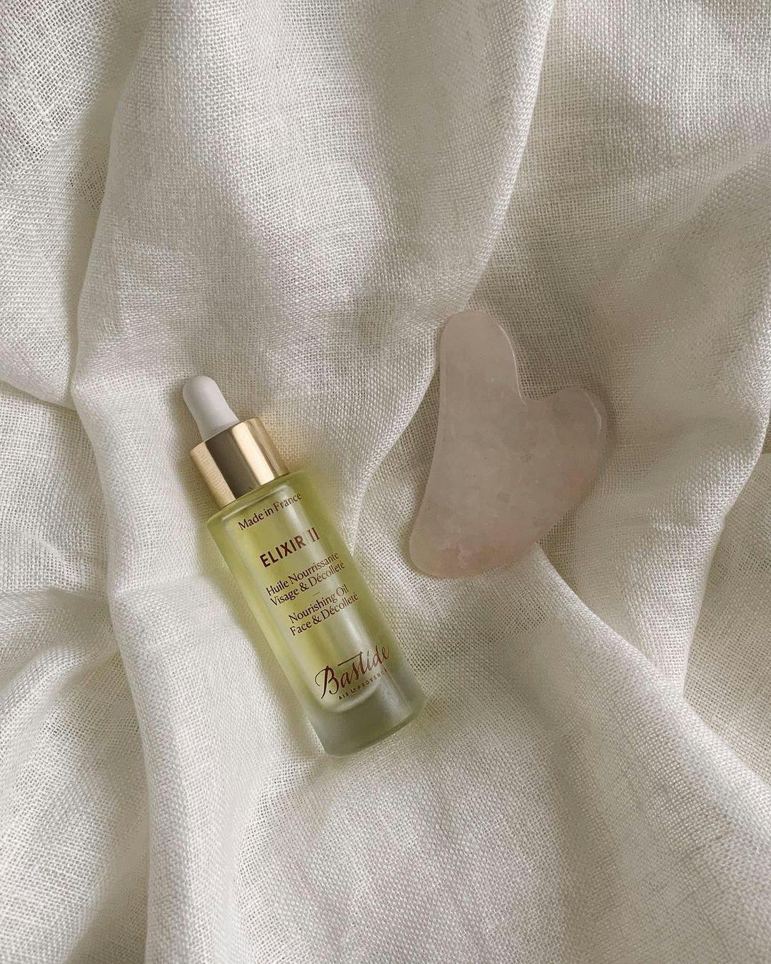 A serum and gua sha lie on a white comforter or sheet.