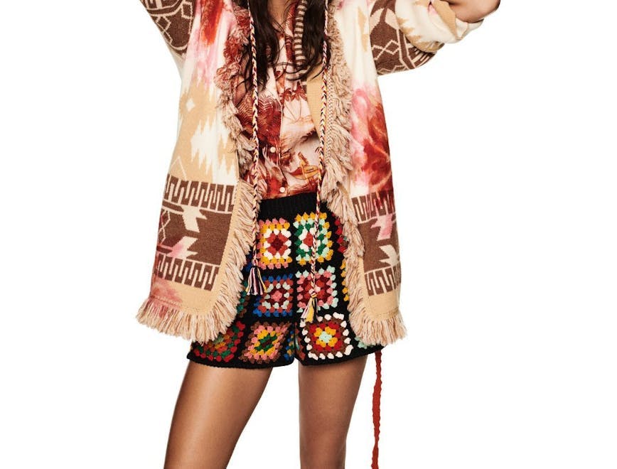 Model wearing crochet shorts and hat, a tie dye shirt and a printed cardigan.