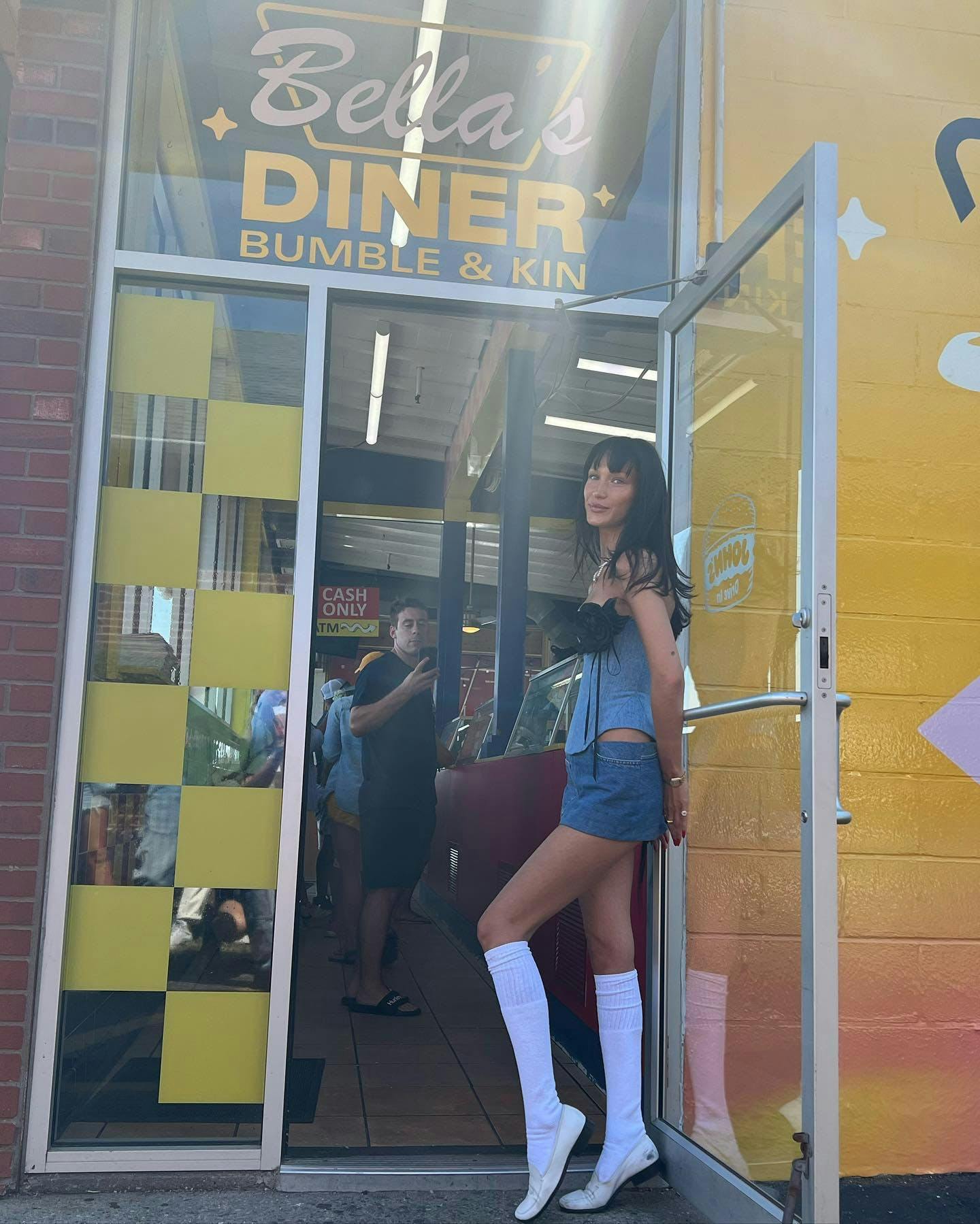 Bella Hadid standing in the doorway of a restaurant called Bella's Diner. She is wearing a denim corset and mini skirt with knee high socks and loafers.