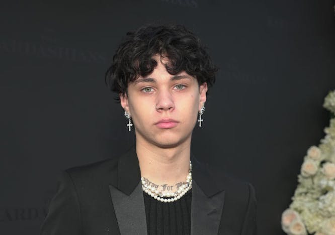 A guy waring a black blazer, black shirt, black pants, pearl necklaces, and cross earrings.