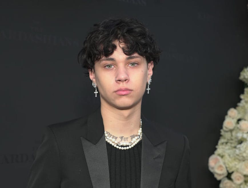A guy waring a black blazer, black shirt, black pants, pearl necklaces, and cross earrings.