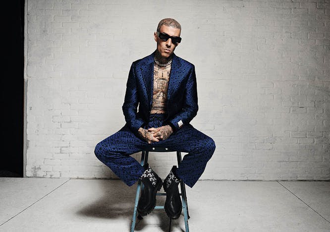 Travis Barker sitting on a stool in a dark blue suit jacket with matching pants.