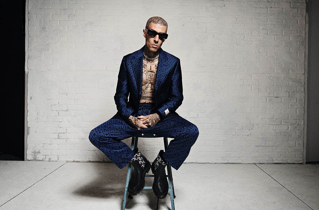 Travis Barker sitting on a stool in a dark blue suit jacket with matching pants.