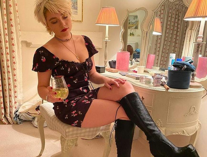 Florence Pugh with short blonde hair and high platform black boots holding a wine glass