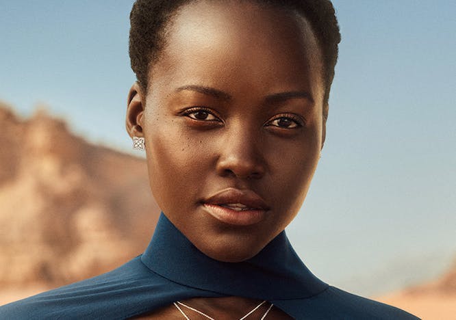 image of lupita nyongo wearing a blue cape and dress wearing silver necklaces, rings, and a bracelet