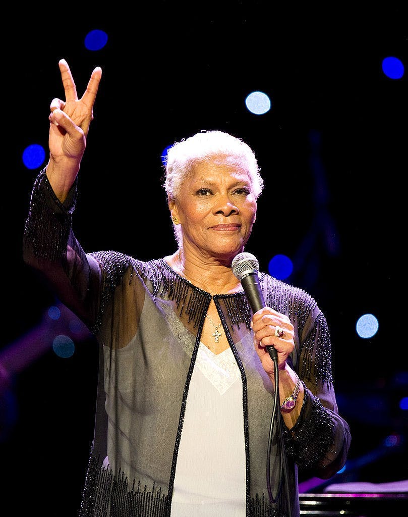 A woman wearing a black sheer jacket and white top while holding a peace sign.