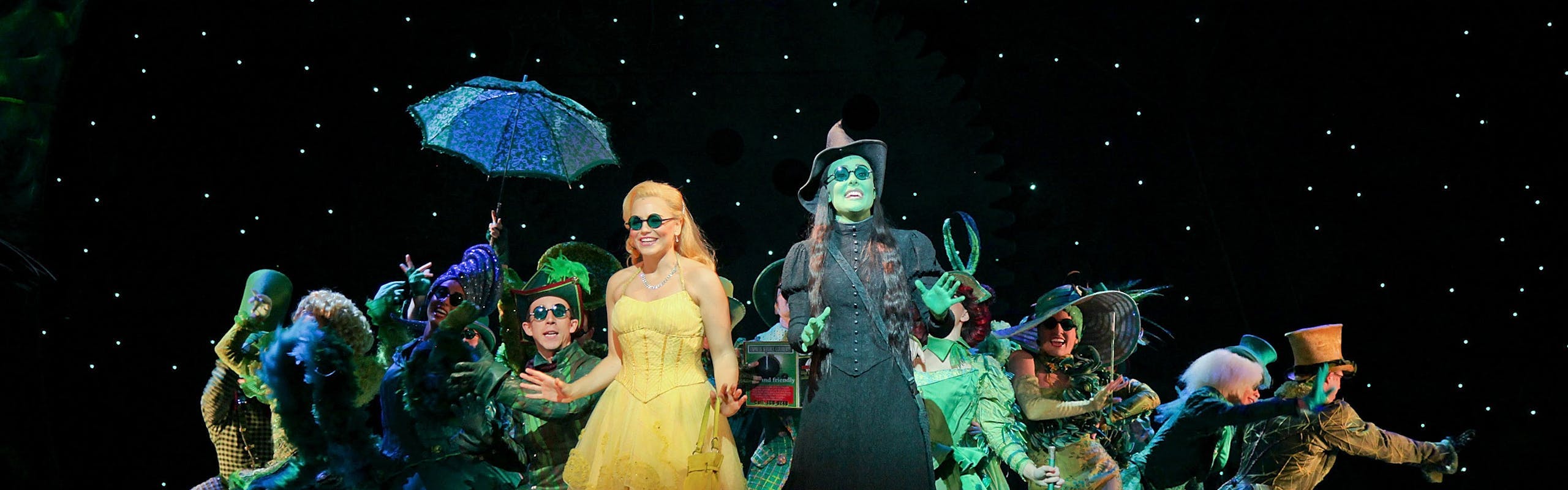 Wicked cast on stage in green Glinda in yellow dress black backdrop