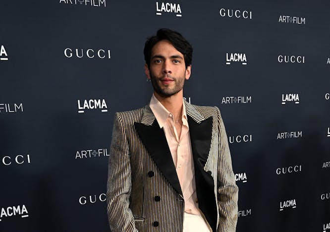 Diego Calva in a brown suit, white pants, on a black and blue background.
