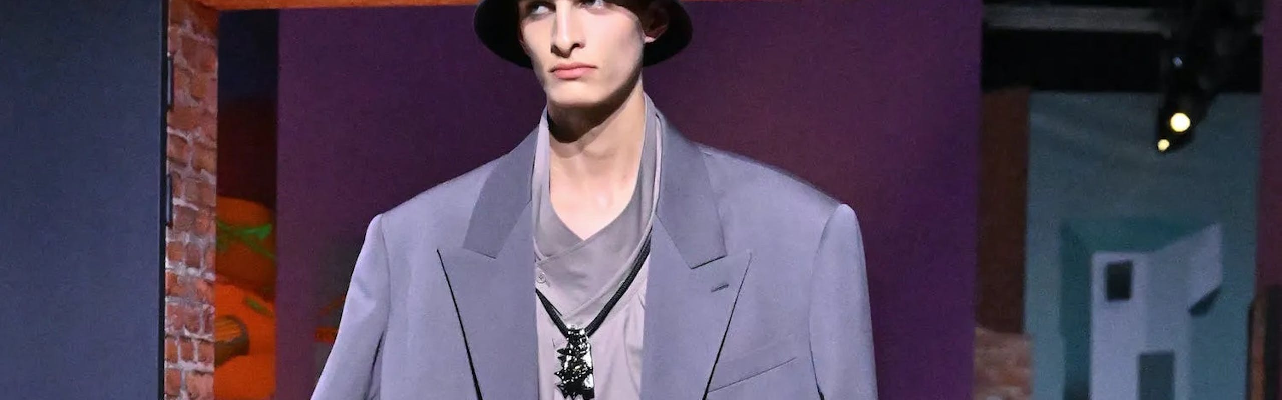 A model in a grey blazer, light purple blouse, and loose grey pants.