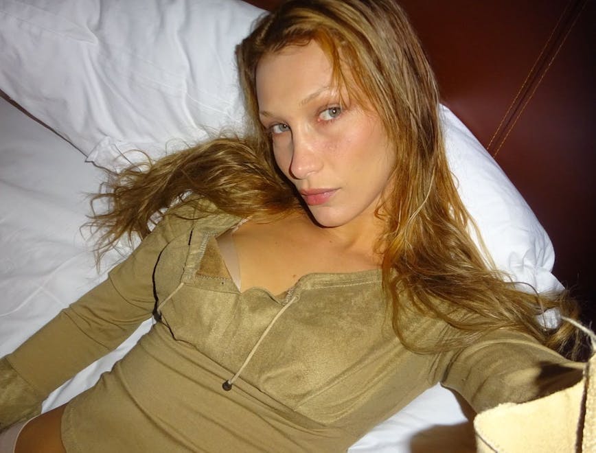 Bella Hadid in green top laying on bed with fresh skin and blonde hair.