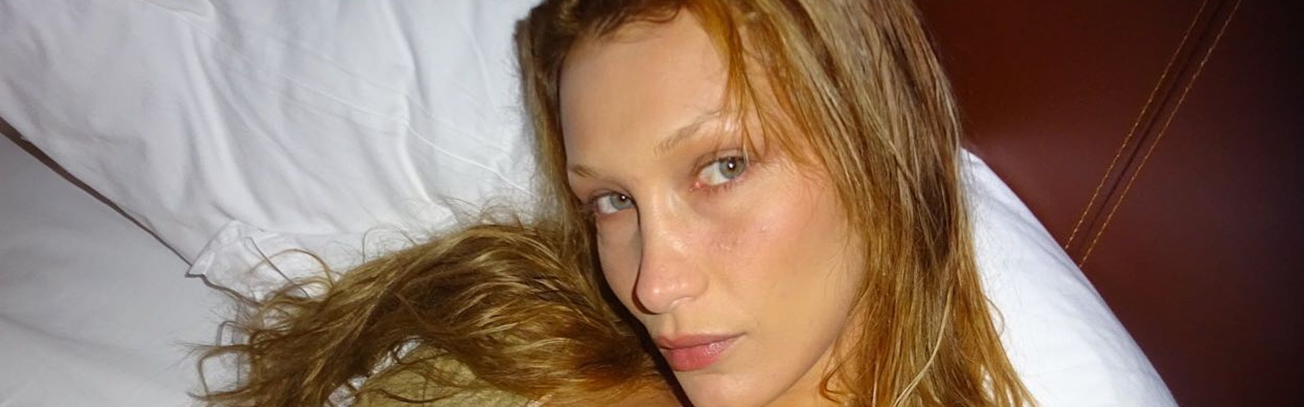 Bella Hadid in green top laying on bed with fresh skin and blonde hair.