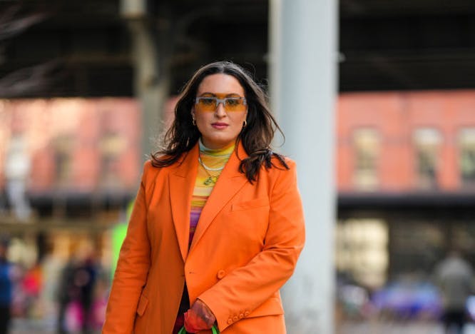 A woman in an orange blazer, multicolored dress, and black boots.