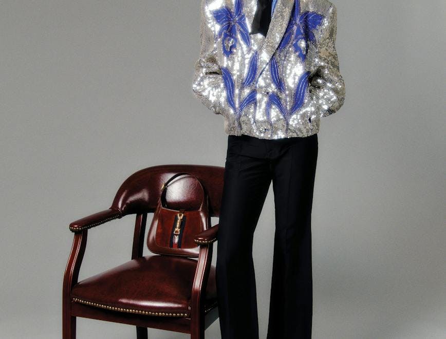 A model in a silver and blue sequined blazer and black pants next to a brown chair.