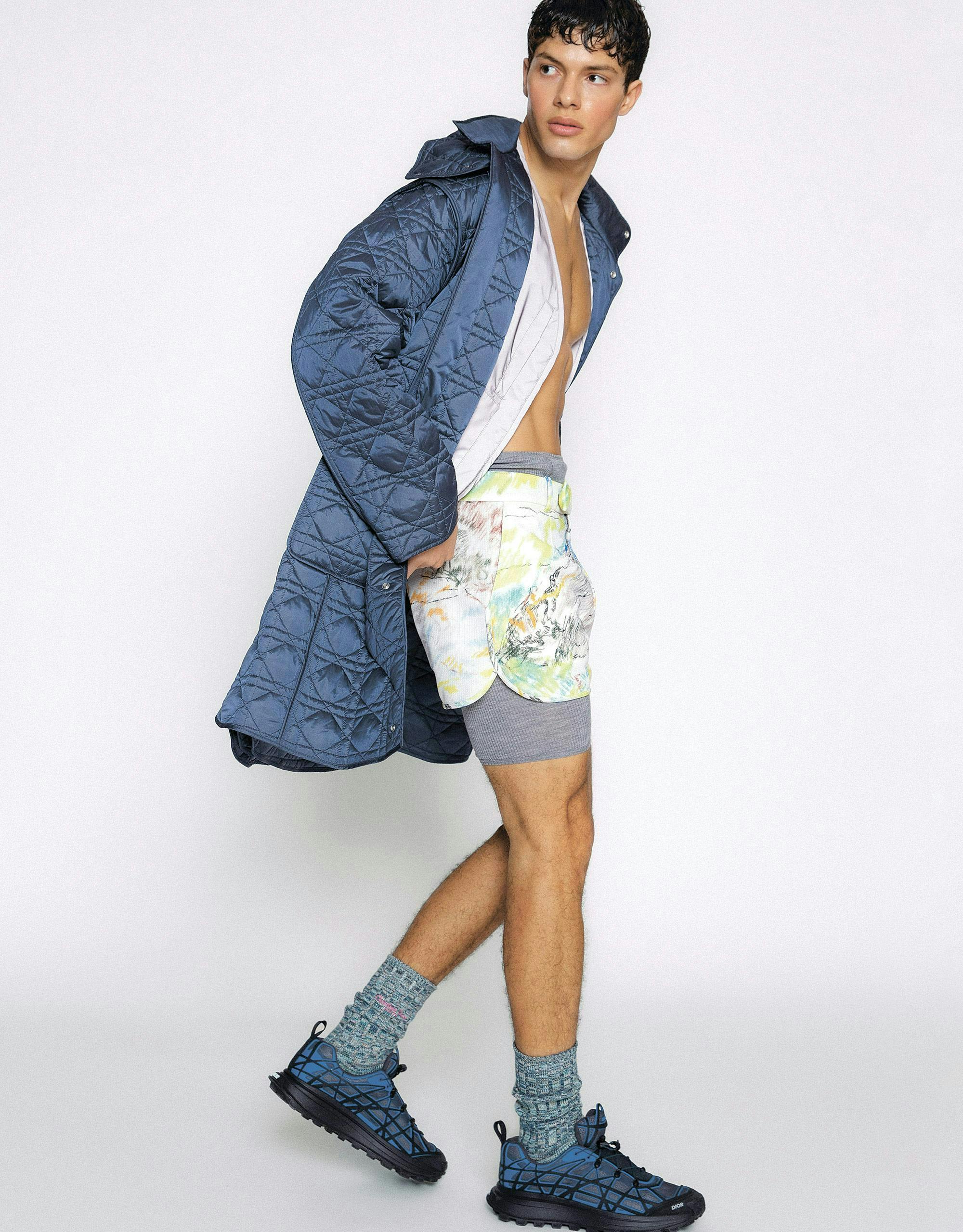 Man wearing blue quilted coat, unbuttoned shirt, shorts, and blue athletic shoes.