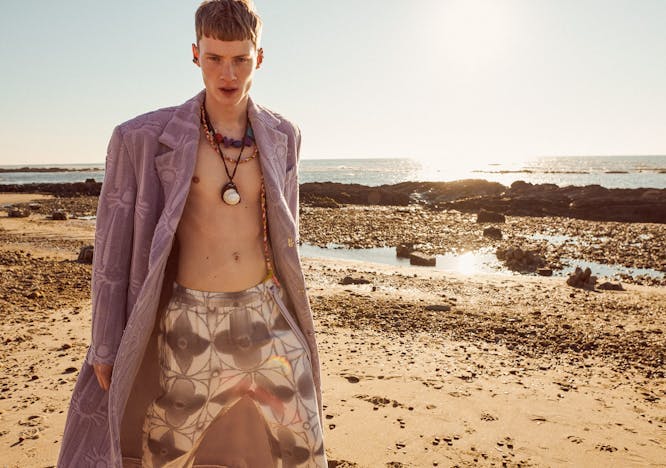 man on the beach wearing purple coat, white and gray printed pants, and multiple necklaces