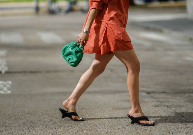 A woman walking in an orange dress and black thong sandals.