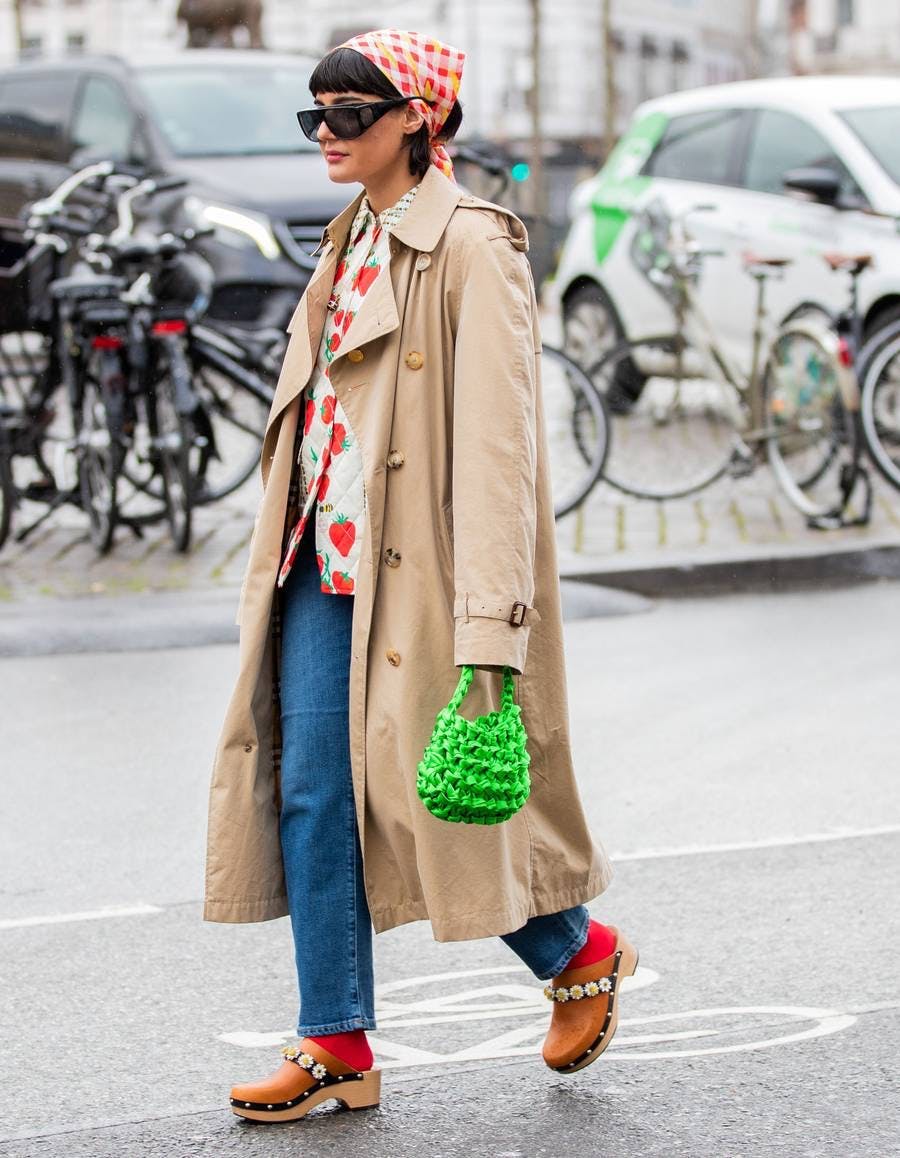 a woman in a trench coat, printed shirt, jeans, and clogs